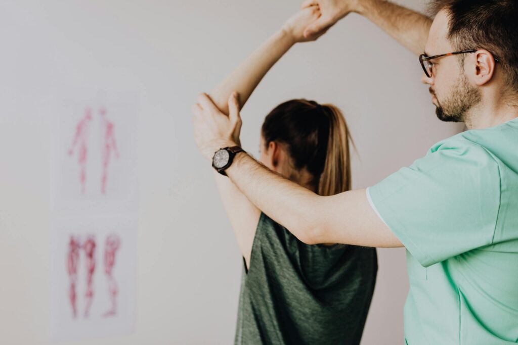 Bearded chiropractor in eyeglasses and wristwatch examining arm of anonymous female in casual clothes with raised hand in doctor office in hospital